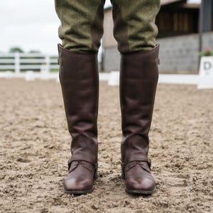 May include: A pair of dark brown leather tall riding gaiters worn over olive green breeches. The gaiters feature a textured leather finish, a side snap button closure at the top, and a buckled strap across the front of the ankle area.