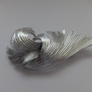 Puede incluir: Broche plateado con un diseño fluido y texturizado. La broche presenta una serie de líneas curvas y paralelas que crean un patrón ondulado. El metal tiene un acabado pulido, reflejando la luz. Un accesorio decorativo para añadir a la ropa.
