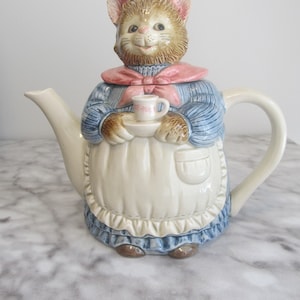 Op de afbeelding: Een keramische theepot in de vorm van een kat die een blauwe jurk en een roze schort draagt, met een klein theekopje in de hand. De theepot heeft een bruin en wit kattengezicht met roze oren en een roze strik.
