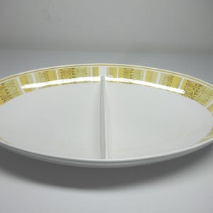 Puede incluir: Un plato ovalado blanco para servir con un centro dividido. El plato tiene un patrón geométrico dorado y amarillo alrededor del borde.