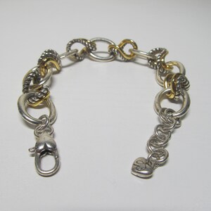 Op de afbeelding: Zilverkleurige armband met goudkleurige accenten. De armband heeft verschillende schakelontwerpen, waaronder ovale, gedraaide en cirkelvormige vormen. Een hartvormige sluiting zet de armband vast. Het is een sieraad.