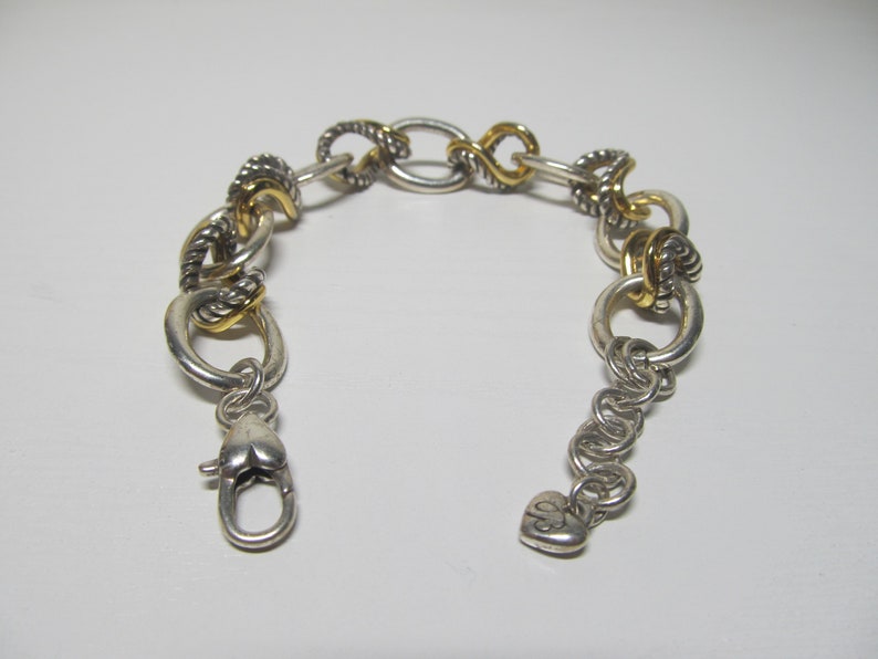 Op de afbeelding: Zilverkleurige armband met goudkleurige accenten. De armband heeft verschillende schakelontwerpen, waaronder ovale, gedraaide en cirkelvormige vormen. Een hartvormige sluiting zet de armband vast. Het is een sieraad.