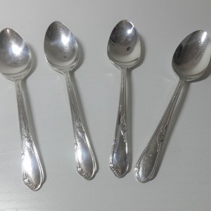 May include: Four silver spoons with oval bowls and ornate floral detailing on the handles. The spoons are arranged on a white surface, reflecting light. A classic silverware set.