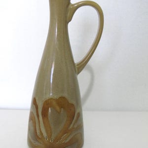 May include: A tan ceramic jug with a curved handle. The jug has a flared base and a narrow neck. A brown floral design decorates the lower portion of the jug. The jug is likely used for serving beverages.