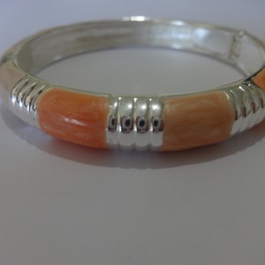 Op de afbeelding: Zilverkleurige scharnierende armband met oranje en perzik emaille segmenten. De armband heeft verticale zilveren banden tussen de emaille secties. Een modieus accessoire. Een stijlvol sieraad.