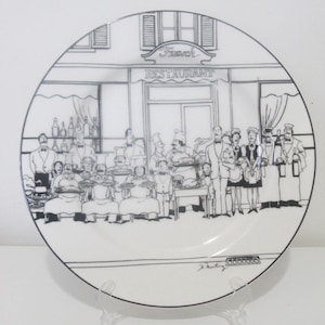 Op de afbeelding: Een wit keramiek bord met een zwart-witte illustratie van een Franse restaurantscène. Het bord toont een winkelgevel met de woorden "French Restaurant" boven de ingang. De scène toont een druk restaurant met klanten die aan tafels zitten en een ober die eten serveert. Het bord is onderaan rechts gesigneerd met "J. Hunting".