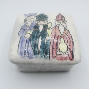 May include: A white ceramic box with a lid, decorated with a colorful illustration of three figures in robes and hats. The figures are holding gifts and appear to be wise men.