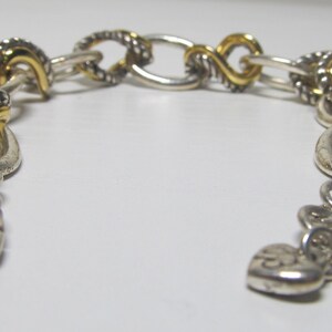 Op de afbeelding: Zilverkleurige armband met goudkleurige accenten. De armband heeft een mix van schakelstijlen, waaronder gedraaide touw- en ovale schakels. Een hartvormige bedel en een karabijnsluiting maken het ontwerp compleet. De armband is ongeveer 18 cm lang.