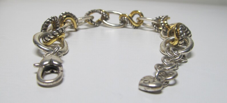 Op de afbeelding: Zilverkleurige armband met goudkleurige accenten. De armband heeft een mix van schakelstijlen, waaronder gedraaide touw- en ovale schakels. Een hartvormige bedel en een karabijnsluiting maken het ontwerp compleet. De armband is ongeveer 18 cm lang.