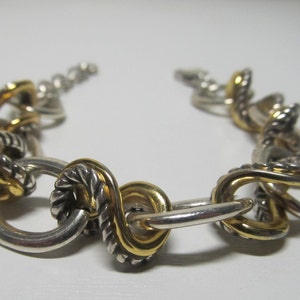 Op de afbeelding: Zilver- en goudkleurige armband met een reeks onderling verbonden schakels. De armband heeft een mix van zilveren en goudkleurige elementen, waarbij sommige schakels een gedraaid touwontwerp hebben. De sluiting is zichtbaar.