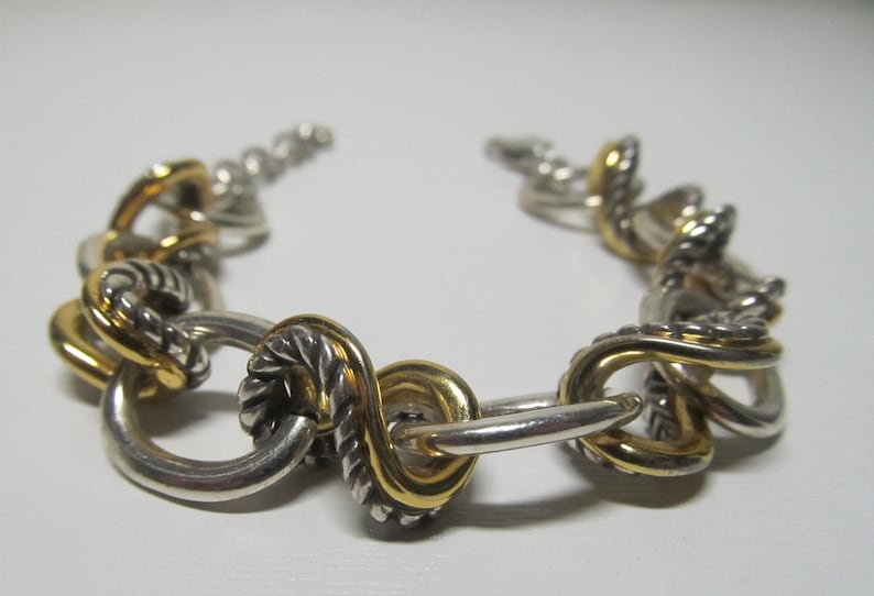 Op de afbeelding: Zilver- en goudkleurige armband met een reeks onderling verbonden schakels. De armband heeft een mix van zilveren en goudkleurige elementen, waarbij sommige schakels een gedraaid touwontwerp hebben. De sluiting is zichtbaar.