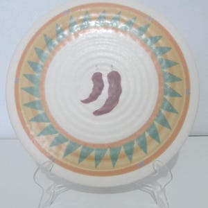 May include: White ceramic plate with a Southwestern design. The plate features a central image of two purple chili peppers. A ring of triangles in teal and yellow surrounds a peach-colored band near the edge.