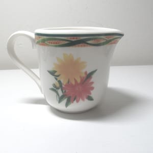 Puede incluir: Crema de cerámica blanca con un diseño floral verde, naranja y amarillo. La crema tiene un asa y una forma redondeada.
