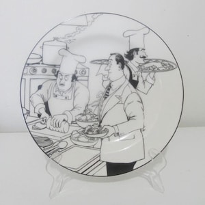 Op de afbeelding: Zwart-witte illustratie van drie koks in een keuken. Een kok houdt een bord met eten vast, een ander rolt deeg en de derde houdt een bord met salade vast. De illustratie bevindt zich op een wit keramiek bord.