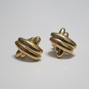 May include: A pair of gold-coloured stud earrings. Each earring has a woven design, with multiple rounded bars crossing over each other. The earrings are set against a white background.