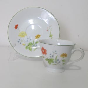 Puede incluir: Un juego de taza y platillo de té blanco con un borde verde y un patrón floral. Las flores son rojas, amarillas y naranjas con hojas verdes y detalles azules.