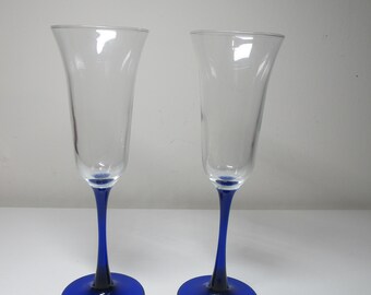 Puede incluir: Dos copas de champán de vidrio transparente con tallos y bases azules. Las copas están vacías y se encuentran sobre una superficie blanca.