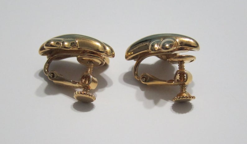May include: A pair of gold-coloured clip-on earrings. Each earring has a rounded, beetle-shaped design with a dark green enamel centre. The clip-on mechanism includes a screw adjustment for a secure fit.