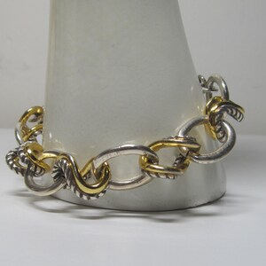 Op de afbeelding: Zilverkleurige armband met goudkleurige accenten. De armband heeft een schakelontwerp met ovale en gedraaide, touwachtige schakels. Goudkleurige schakels zijn door de armband verspreid, wat een vleugje contrast en elegantie aan de sieraden toevoegt.