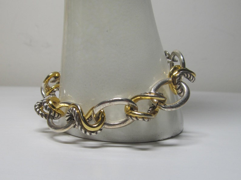 Op de afbeelding: Zilverkleurige armband met goudkleurige accenten. De armband heeft een schakelontwerp met ovale en gedraaide, touwachtige schakels. Goudkleurige schakels zijn door de armband verspreid, wat een vleugje contrast en elegantie aan de sieraden toevoegt.