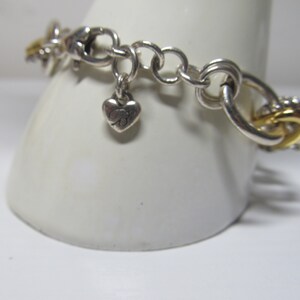 Op de afbeelding: Zilverkleurige armband met gouden accenten. De armband heeft een hartvormige bedel met de letter "B" en een mix van ronde en in elkaar grijpende schakels. Goudkleurig metaal wordt gebruikt in de schakels en decoratieve elementen.