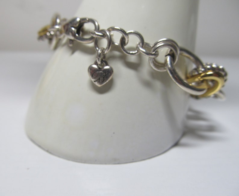 Op de afbeelding: Zilverkleurige armband met gouden accenten. De armband heeft een hartvormige bedel met de letter "B" en een mix van ronde en in elkaar grijpende schakels. Goudkleurig metaal wordt gebruikt in de schakels en decoratieve elementen.