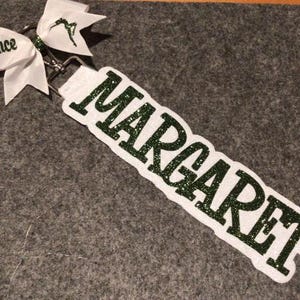 May include: A white fabric keychain featuring the name MARGARET in dark green glitter lettering. The keychain is attached to a silver metal lobster clasp and a white ribbon bow with the text dance and a small green figure printed on it.