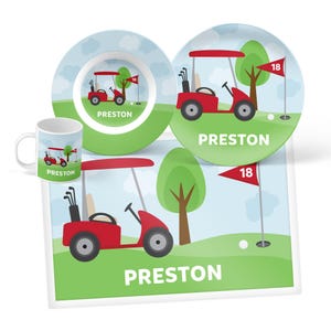 May include: A dining set featuring a mug, a bowl, and a plate, each decorated with a red golf cart, a green golf course, a tree, and a red flag labeled 18. The name PRESTON is printed in white capital letters on each item in the set.