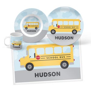 May include: A dining set featuring a mug, a small bowl, a plate, and a rectangular placemat. Each item displays a yellow school bus graphic, a light blue sky with white clouds, and the name HUDSON printed in dark gray capital letters.