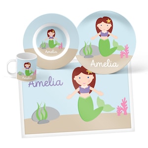 May include: A blue and brown children's dinnerware set with a mermaid theme. The set includes a bowl, plate, mug, and placemat. The placemat features a mermaid with green hair and tail, and the text "Amelia".