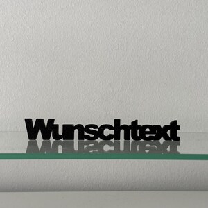 Könnte beinhalten: Ein schwarzer, dreidimensionaler Schriftzug mit dem Wort "Wunschtext" in einer fetten, serifenlosen Schriftart. Die Buchstaben stehen auf einer klaren, horizontalen Glasfläche, wodurch sich das Wort direkt unter dem Objekt spiegelt.