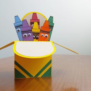 Puede incluir: Una caja amarilla con rayas verdes se abre para revelar una tarjeta emergente con lápices de colores con ojos saltones. Los lápices están dispuestos en un círculo.