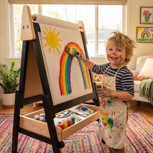 Könnte beinhalten: Eine hölzerne Staffelei mit schwarzem Rahmen und einer Ablage aus Naturholz. Ein Kind malt einen Regenbogen und eine Sonne auf das Papier. Das Kind trägt ein gestreiftes Hemd und eine mit Farbe bespritzte Schürze. Malutensilien befinden sich in der Ablage.