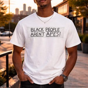 May include: White t-shirt with the text "BLACK PEOPLE AREN'T APES!" in a black, bold font. The shirt is worn by a person standing outdoors. The person is wearing black jeans and a silver necklace and watch.