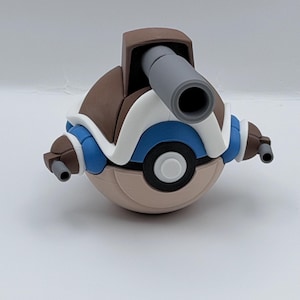 May include: A spherical toy figure featuring a brown and white shell-like top with a large gray cannon protruding from the center. The base is a tan sphere with a central black and white circular button. Two small gray cannons extend from the sides.