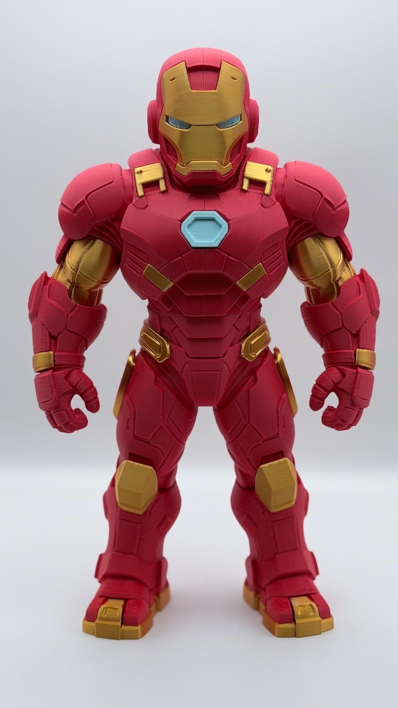 May include: A standing Iron Man action figure featuring a matte red and metallic gold armored suit. The character has a light blue hexagonal chest piece, a gold faceplate, and a robust, segmented design with visible 3D printed texture lines.
