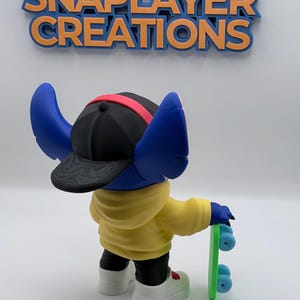 May include: A blue character figurine wearing a black cap with a red band, yellow hoodie, black pants, and white shoes, holding a green skateboard. The figurine has large blue ears. The text "FOLLOW SNAPPLAYER CREATIONS" is above the figurine.