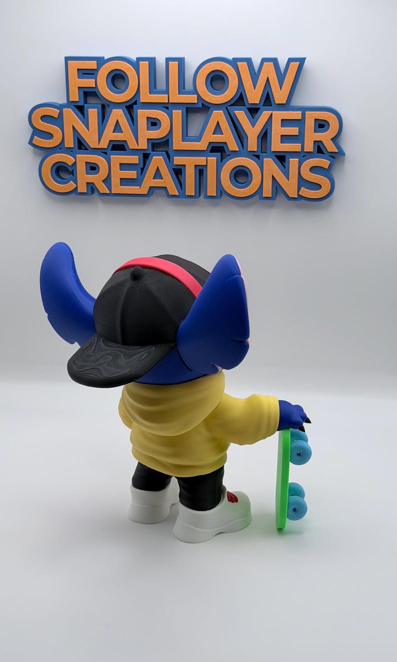 May include: A blue character figurine wearing a black cap with a red band, yellow hoodie, black pants, and white shoes, holding a green skateboard. The figurine has large blue ears. The text "FOLLOW SNAPPLAYER CREATIONS" is above the figurine.
