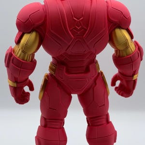 May include: A rear view of a red and gold robotic action figure. The figure features a matte red finish on the armor plating with metallic gold accents on the upper arms, waist, and feet. The design includes segmented armor panels.