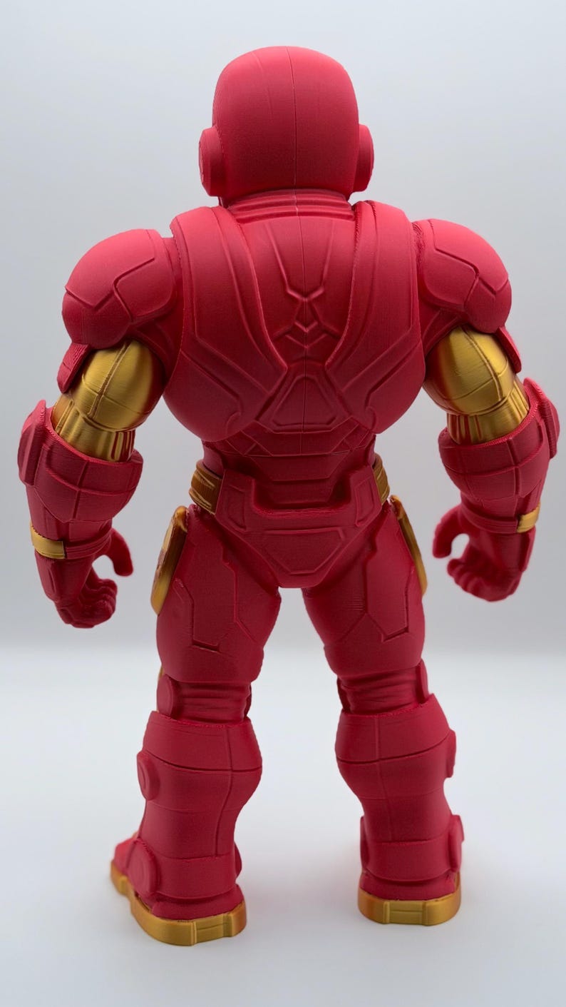 May include: A rear view of a red and gold robotic action figure. The figure features a matte red finish on the armor plating with metallic gold accents on the upper arms, waist, and feet. The design includes segmented armor panels.