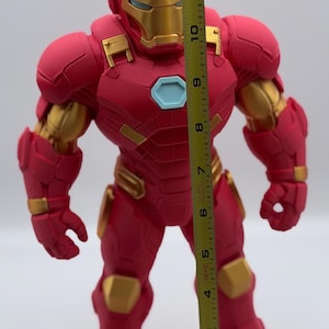 May include: A red and gold Iron Man action figure standing next to a yellow measuring tape. The tape measure indicates the figure stands approximately 12.5 inches tall. The figure features a light blue chest arc reactor and detailed armor plating.