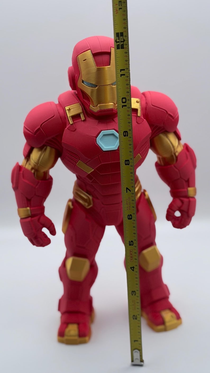 May include: A red and gold Iron Man action figure standing next to a yellow measuring tape. The tape measure indicates the figure stands approximately 12.5 inches tall. The figure features a light blue chest arc reactor and detailed armor plating.