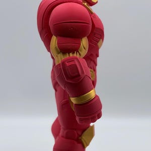 May include: A side view of a red and gold armored action figure. The figure features a helmet with a gold faceplate, segmented shoulder armor, and a bulky, mechanical design with visible 3D printed layer lines throughout the red surface.