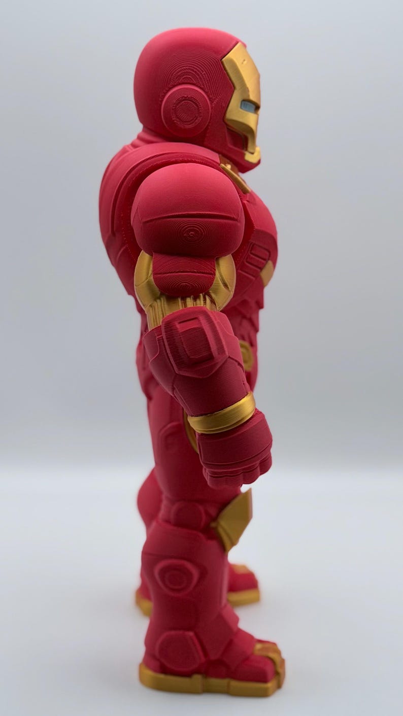 May include: A side view of a red and gold armored action figure. The figure features a helmet with a gold faceplate, segmented shoulder armor, and a bulky, mechanical design with visible 3D printed layer lines throughout the red surface.