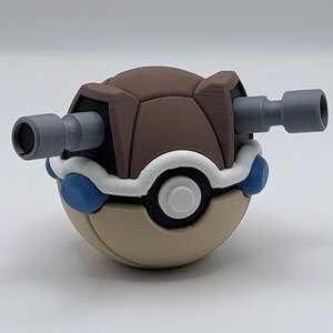 May include: A 3D-printed, multi-colored toy resembling a Pokémon character. The toy features a brown shell, a tan base, white and blue accents, and two gray, cylindrical cannons extending from the sides. The design is reminiscent of a turtle.