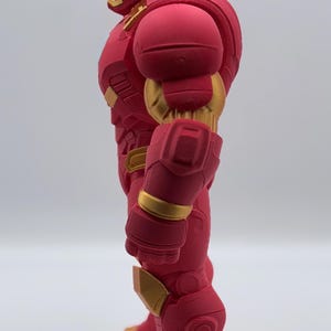 May include: A side profile view of a stylized figurine of a character wearing a red and gold armored suit. The figure features a smooth, matte red finish on the main body panels with contrasting metallic gold accents on the face, arms, and feet.