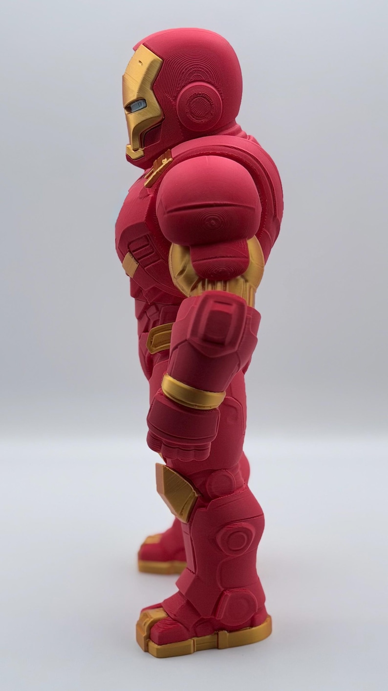 May include: A side profile view of a stylized figurine of a character wearing a red and gold armored suit. The figure features a smooth, matte red finish on the main body panels with contrasting metallic gold accents on the face, arms, and feet.