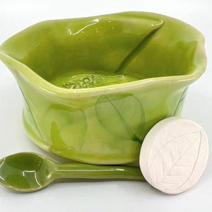May include: A green ceramic bowl with a leaf design, a matching spoon, and a white, leaf-imprinted soap. The bowl has a unique shape and the set is likely for serving or display. The spoon is approximately 5 inches long.