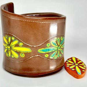 May include: A brown ceramic cup with a curved handle and a matching coaster. The cup features a yellow and teal floral design. The coaster has a similar floral pattern. The cup is approximately 4 inches tall.
