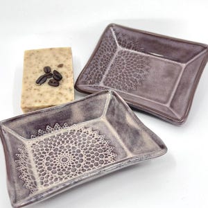 May include: Two ceramic soap dishes with a decorative lace pattern in a muted purple glaze. A bar of soap with coffee beans is also present. The dishes have a rectangular shape with slightly curved sides.